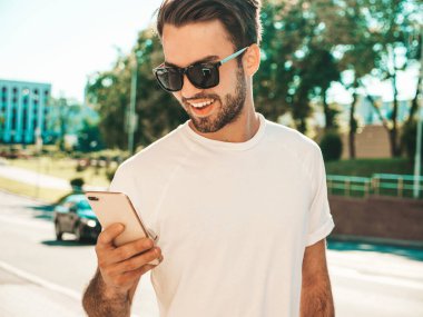 Yakışıklı, şık, havalı, lamberseksüel bir model. Beyaz tişört giymiş bir adam. Sokakta poz veren bir erkek. Cep telefonunda tutuyor, kaydırıyor ve mesaj atıyor. Akıllı telefondan mesaj yazar