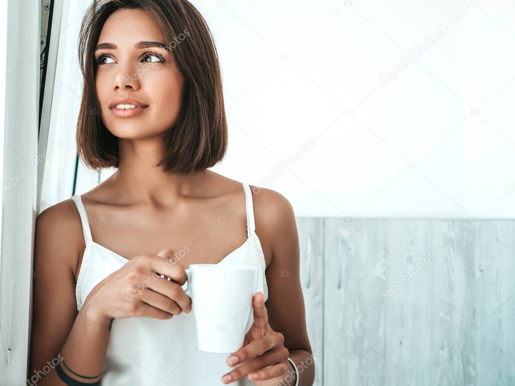Portrait of beautiful girl dressed in white pajamas. Carefree woman holds cup and drinking coffee. Sexy model enjoying her morning breakfast at balcony. Female in her thoughts 