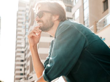 Yakışıklı, gülümseyen stil sahibi Lambersex modelinin portresi. Yeşil tişörtlü adam. Moda erkek gökdelen arka planında güneş gözlükleriyle sokakta poz veriyor. Gün batımında dışarıda. 