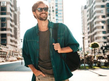 Yakışıklı, gülümseyen stil sahibi Lambersex modelinin portresi. Yeşil tişörtlü adam. Moda erkek gökdelen arka planında güneş gözlükleriyle sokakta poz veriyor. Gün batımında dışarıda. 