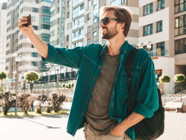 Yakışıklı, gülümseyen stil sahibi Lambersex modelinin portresi. Yeşil tişörtlü adam. Moda erkek gökdelen arka planında güneş gözlükleriyle sokakta poz veriyor. Gün batımında dışarıda. Selfie çekiyorum.
