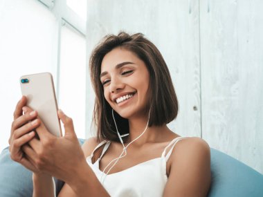 Beyaz pijama giymiş ve kulaklıkla müzik dinleyen güzel bir kadının portresi. Yumuşak koltukta oturan tasasız manken. Seksi kadın balkonda eğleniyor. 
