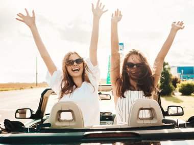 Üstü açık arabadaki iki güzel ve gülümseyen hippi kadının portresi. Cabriolet kullanan kaygısız seksi kadın. Pozitif modeller güneş gözlükleriyle geziyor ve eğleniyorlar. El kaldırıyorlar.