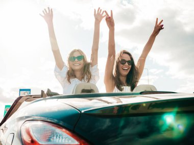 Üstü açık arabadaki iki güzel ve gülümseyen hippi kadının portresi. Cabriolet kullanan kaygısız seksi kadın. Pozitif modeller güneş gözlükleriyle geziyor ve eğleniyorlar. El kaldırıyorlar.