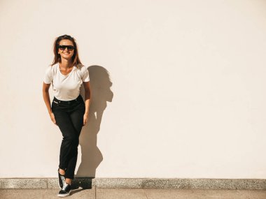 Güneş gözlüklü güzel bir modelin portresi. Hipster beyaz tişört ve kot pantolon giymiş bir kadın. Sokakta duvar kenarında poz veren moda bir kadın.