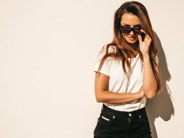 Güneş gözlüklü güzel bir modelin portresi. Hipster beyaz tişört ve kot pantolon giymiş bir kadın. Sokakta duvar kenarında poz veren moda bir kadın.