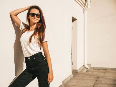 Güneş gözlüklü güzel bir modelin portresi. Hipster beyaz tişört ve kot pantolon giymiş bir kadın. Sokakta duvar kenarında poz veren moda bir kadın.