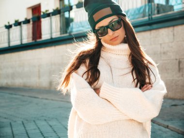Güzel gülümseyen modelin portresi. Sıcak hippi beyaz kazak ve bere giymiş bir kadın. Günbatımında güneş gözlükleriyle sokakta poz veren moda bir kadın.