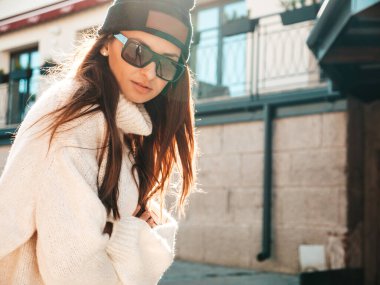 Güzel gülümseyen modelin portresi. Sıcak hippi beyaz kazak ve bere giymiş bir kadın. Günbatımında güneş gözlükleriyle sokakta poz veren moda bir kadın.