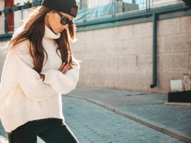 Güzel gülümseyen modelin portresi. Sıcak hippi beyaz kazak ve bere giymiş bir kadın. Günbatımında güneş gözlükleriyle sokakta poz veren moda bir kadın.