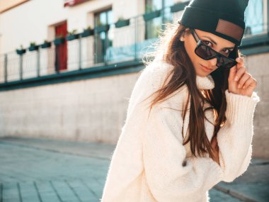 Güzel gülümseyen modelin portresi. Sıcak hippi beyaz kazak ve bere giymiş bir kadın. Günbatımında güneş gözlükleriyle sokakta poz veren moda bir kadın.