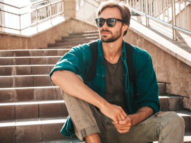 Yakışıklı, şık bir hipster Lambersex modelinin portresi. Yeşil gömlekli bir adam. Moda erkeği, güneş gözlüklü arka planda, merdivenlerde oturuyor.
