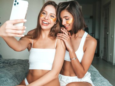 Gözlerinin altında bantlarla gülümseyen iki güzel kadın. Otel odasında yatakta oturan kaygısız seksi mankenler. Evde güzellik tedavisi yapıyorlar ve Instagram için selfie çekiyorlar.