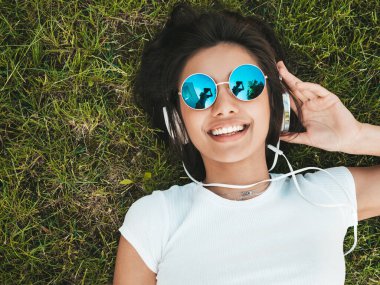Parktaki çimlerin üzerinde uzanan genç ve şık hippi kadının moda portresi. Moda kıyafetleri giyiyor. Hafta sonlarının tadını çıkarıyor. Kadın kulaklıkla müzik dinliyor. Üst görünüm