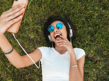 Parktaki çimlerin üzerinde uzanan zarif hippi kadının moda portresi. Moda kıyafetleri giyiyor. Selfie yapıyor. Kadın kulaklıkla müzik dinliyor. Üst görünüm