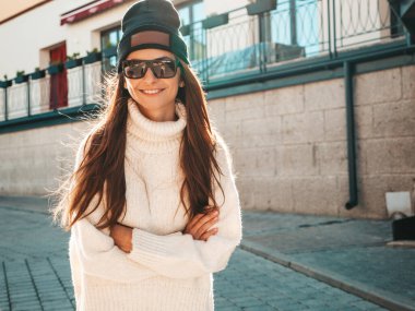 Güzel gülümseyen modelin portresi. Sıcak beyaz kazak ve bere giymiş bir kadın. Gün batımında güneş gözlükleriyle sokakta poz veriyor.