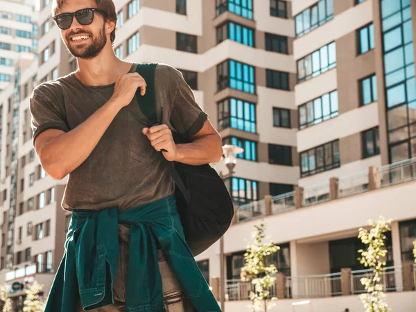 Yakışıklı, gülümseyen stil sahibi Lambersex modelinin portresi. Yeşil tişörtlü adam. Moda erkek gökdelen arka planında güneş gözlükleriyle sokakta poz veriyor. Gün batımında sırt çantasıyla dışarıda.