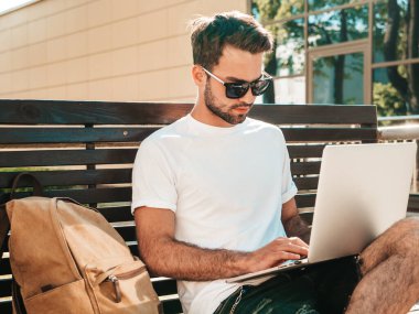 Yakışıklı, güler yüzlü, stil sahibi hipster Lambersex modeli. Moda adamı sokakta bankta oturuyor ve dizüstü bilgisayar kullanıyor, mesaj yazıyor. Ortak çalışma konsepti. Teknoloji ve iletişim