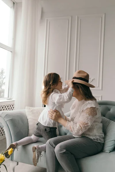 Young mom and little smiling preschooler daughter sit on couch in living room sharing secrets gossiping, mother whisper in excited small girl child ear talking chatting enjoy close communication