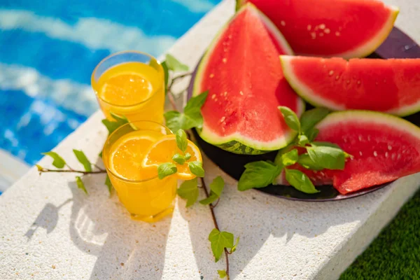 Concepto tropical de vacaciones de verano. Zumo de naranja fresco y jugo de sandía y piña en el borde de una piscina . — Foto de Stock