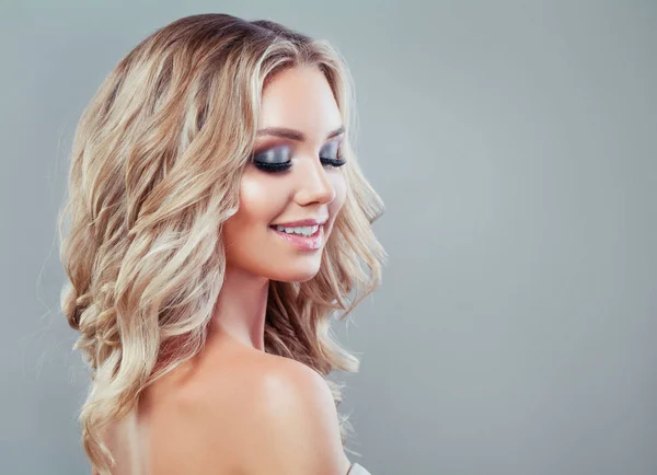 Smiling blonde woman with healthy wavy hair and makeup on blue background