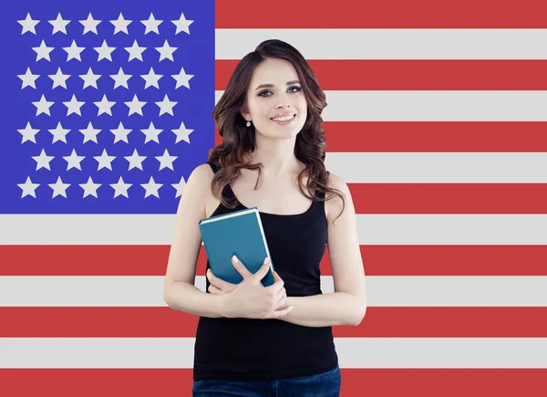 Mujer bonita estudiante sonriendo y sosteniendo el libro contra el fondo de la bandera de los Estados Unidos de América. Viaja a Estados Unidos y aprende inglés americano —  Fotos de Stock