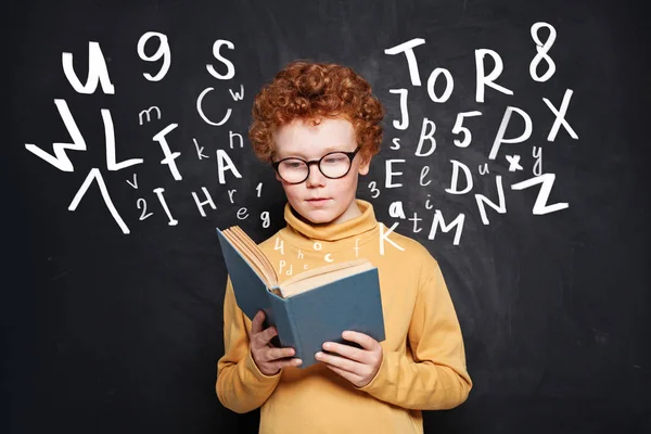 Curioso ragazzino con gli occhiali che legge un libro contro la lavagna — Foto Stock