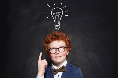 Takım elbiseli küçük çocuk, tahta arka plandaki ampulü işaret ediyor.