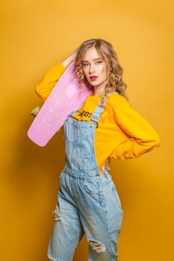 Parlak sarı arka planda pembe kaykay tutan moda kadını.