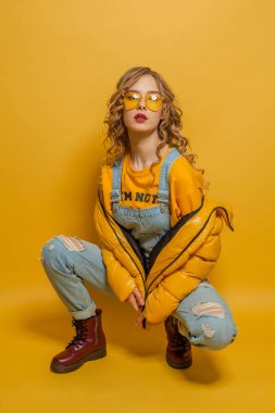 Parlak sarı arka planda poz veren kışlık ceketli manken kadın.
