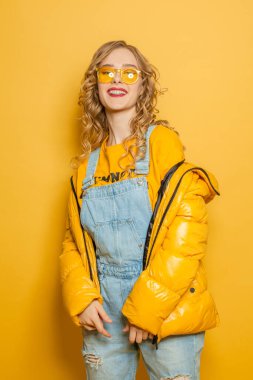 Parlak sarı arka planda duran kışlık ceketli mutlu manken kız.
