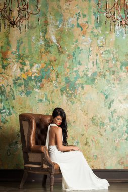 Uzun saçlı ve makyajlı şık bir kadın lüks bir iç mekanda oturuyor.