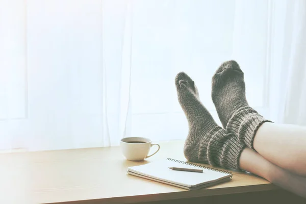 妇女休息保持腿在温暖的袜子桌与早晨咖啡和笔记簿 文本空间 — 图库照片