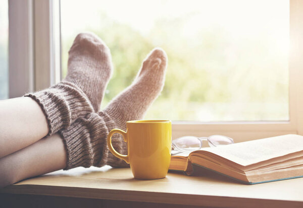Woman resting keeping legs in warm socks on table with morning coffee and reading book