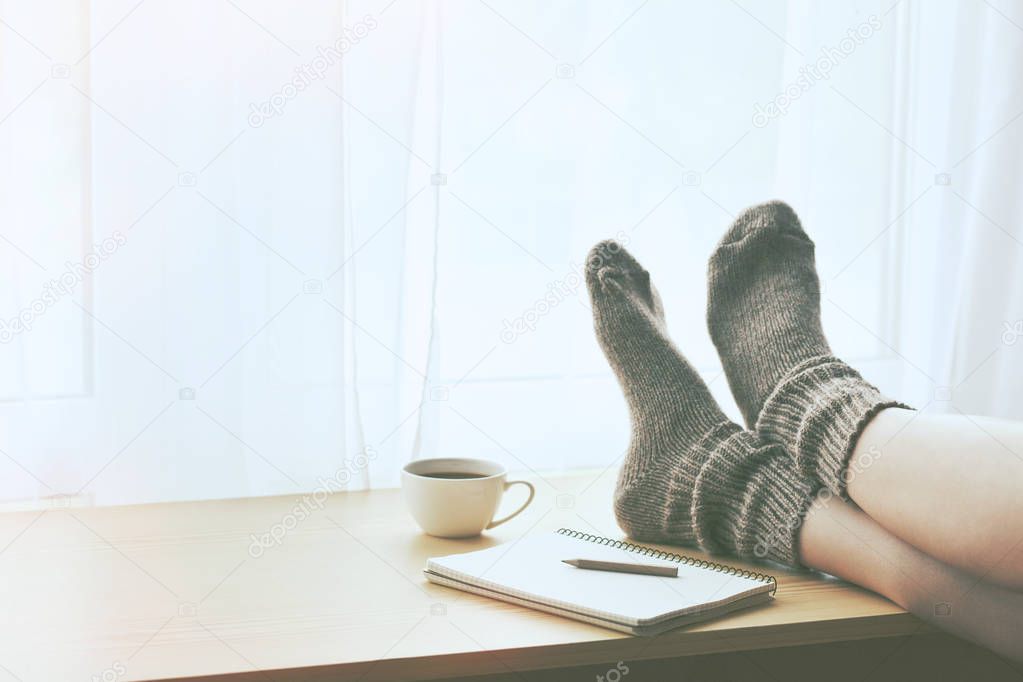 Woman resting keeping legs in warm socks on table with morning coffee and notebook. Space for text