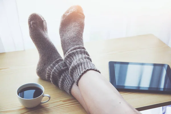 Woman resting keeping legs in warm socks on table with morning coffee and reading e-book or tablet
