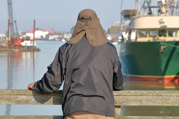 A man wearing head cover that protects against extreme sun exposure, stands on a deck overlooking a boat marina.