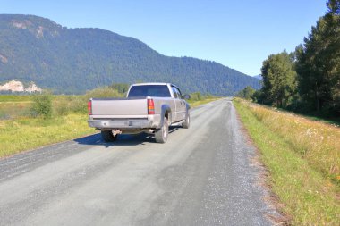 Wide view of a pickup truck as it drives on a long, narrow gravel road embankment in a scenic mountainous valley.  clipart