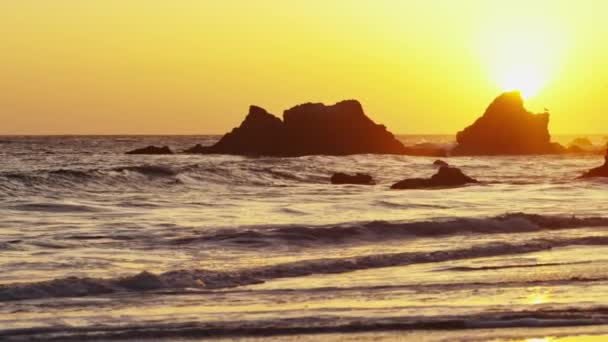 Pequeñas olas del océano golpeando la orilla con el sol poniéndose detrás de una roca . — Vídeo de stock