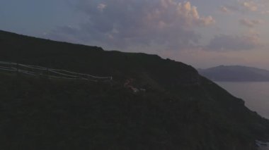 Gün batımında deniz kenarında uçurumlarda oturan çift üzerinde İHA