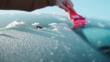 Kırmızı kazıyıcılı bir adam buzu kaldırmak için arabanın ön camını kullanıyor.