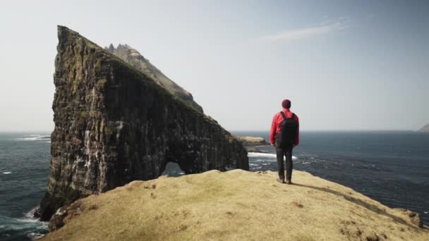 Человек с рюкзаком, стоящим на краю скалы, чтобы полюбоваться скалой Дрангарнира — стоковое видео