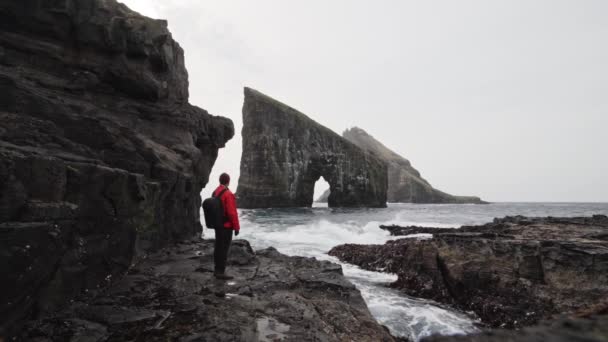 Человек в красной куртке на скалах с видом на Дрангарнирскую арку на Фарерских островах — стоковое видео