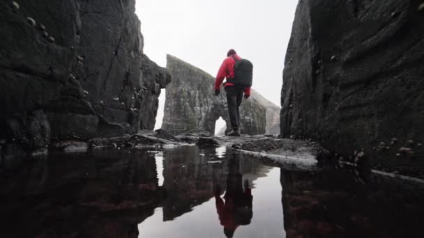 Hombre de chaqueta roja caminando por rocas rocosas y poco profundas en las Islas Feroe — Vídeos de Stock