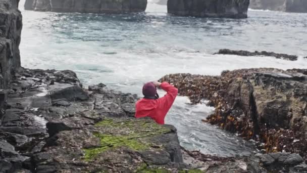 Man i röd jacka sitter på stenar tar en bild av havsvågor i Drangarnir — Stockvideo
