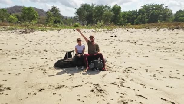 Casal sorrindo e sentado em log com drone puxando para fora revelando praia de areia — Vídeo de Stock