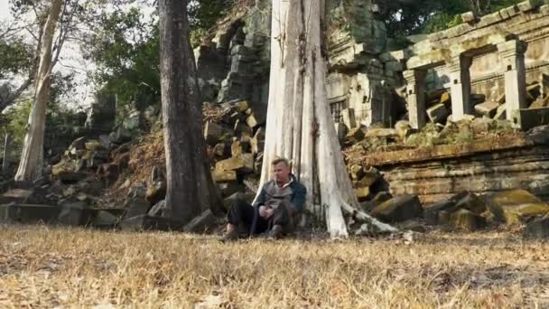 Καμποτζιανά ερείπια ναού και ένας εξερευνητής κάθεται από το παλιό δέντρο — Αρχείο Βίντεο