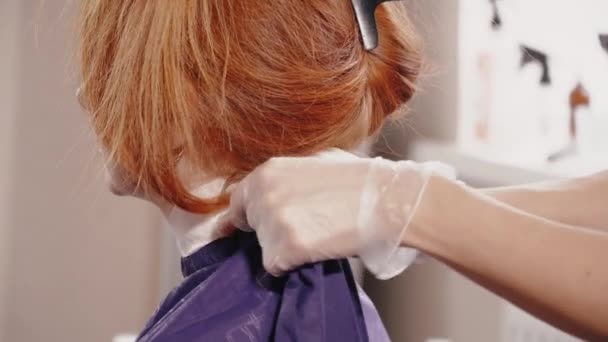 Парикмахер в перчатках закрепляет пеньюар на шее клиента — стоковое видео