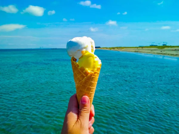 Hand is holding the vanilla ice cream. In the background is blue sky, sea nd sandy beach. — Stock Photo, Image