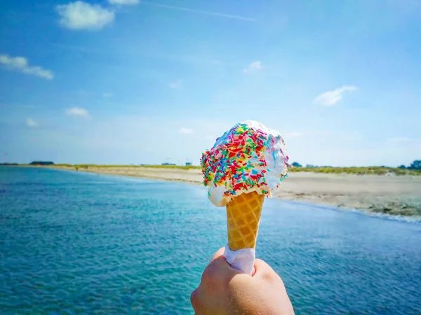 A mão está a segurar o gelado de baunilha. No fundo é o céu azul, mar e praia de areia. — Fotografia de Stock